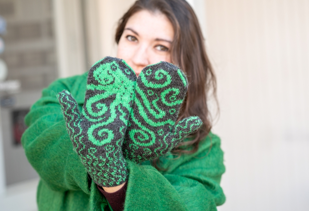 A woman holding her hands up towards the camera, palms out, wrists crossed so the thumbs are on the outside. The octopus on her handknit mittens flows across both palms.